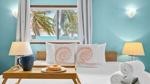 A hotel room with a bed, two pillows with a spiral design, a tray with a mug and pastries, a starfish decoration, and a view of palm trees through the window.