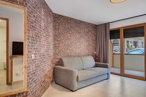 Une pièce avec un mur en briques, un canapé gris, un miroir encadré en bois et une porte-fenêtre coulissante donnant sur un espace extérieur.