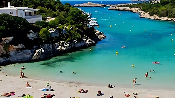 Ein Strand mit klarem türkisfarbenem Wasser, felsiger Küstenlinie und einem weißen Gebäude mit Balkon.