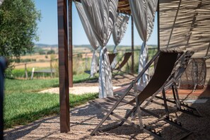 Un espace extérieur aménagé avec des sièges, baigné de lumière naturelle, et offrant une vue sur la campagne, notamment sur un arbre et un champ d’herbe.