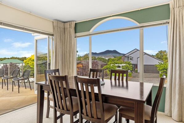 A dining area with a table and chairs, large windows with curtains, and a view of houses and greenery outside.