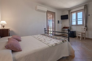 A bedroom with a bed, a bedside table with a lamp, a chair, a desk with a computer, a window with curtains, and an air conditioning unit. - Room 'Giusti - 5 Sughera' with Shared Garden, Wi-Fi and Air Conditioning (Magliano in Toscana)