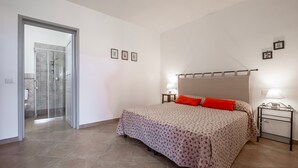 Ein Schlafzimmer mit Bett, Nachttisch, Lampe, Duschbereich und einer Tür zu einem weiteren Raum.