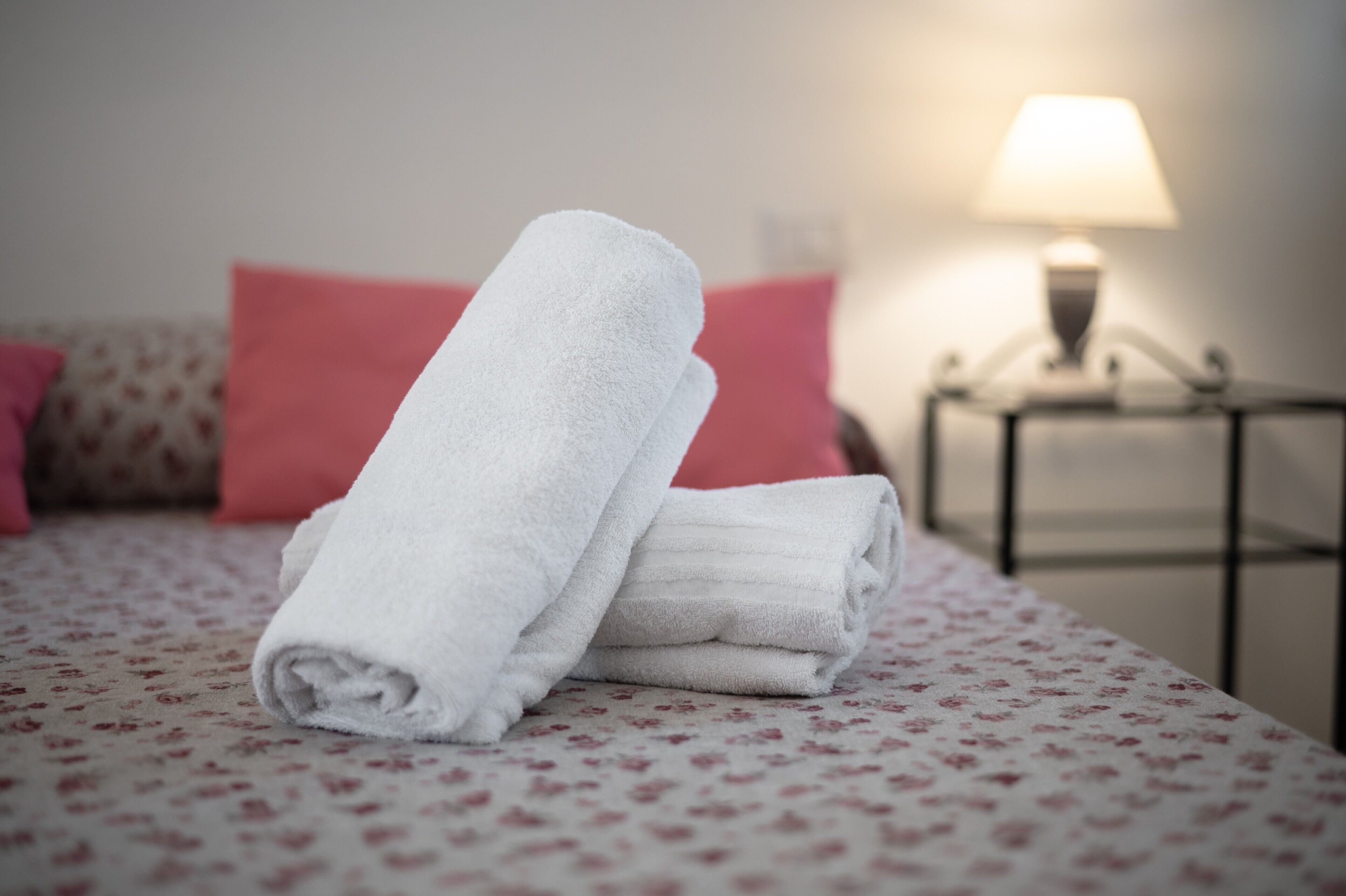 A neatly made bed with rolled white towels, a floral-patterned bedspread, and a lamp on a bedside table.