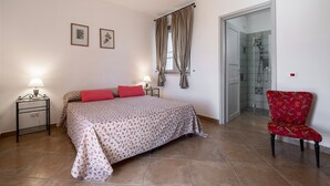 A bedroom with a bed, two bedside lamps, a chair, a shower area, and a door leading to another room. - Room 'Giusti - 3 Leccio' with Shared Garden, Wi-Fi and Air Conditioning (Magliano in Toscana)