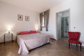 Camera da letto con un letto, due lampade ai lati del letto, una sedia, zona doccia e una porta che dà su un'altra stanza.