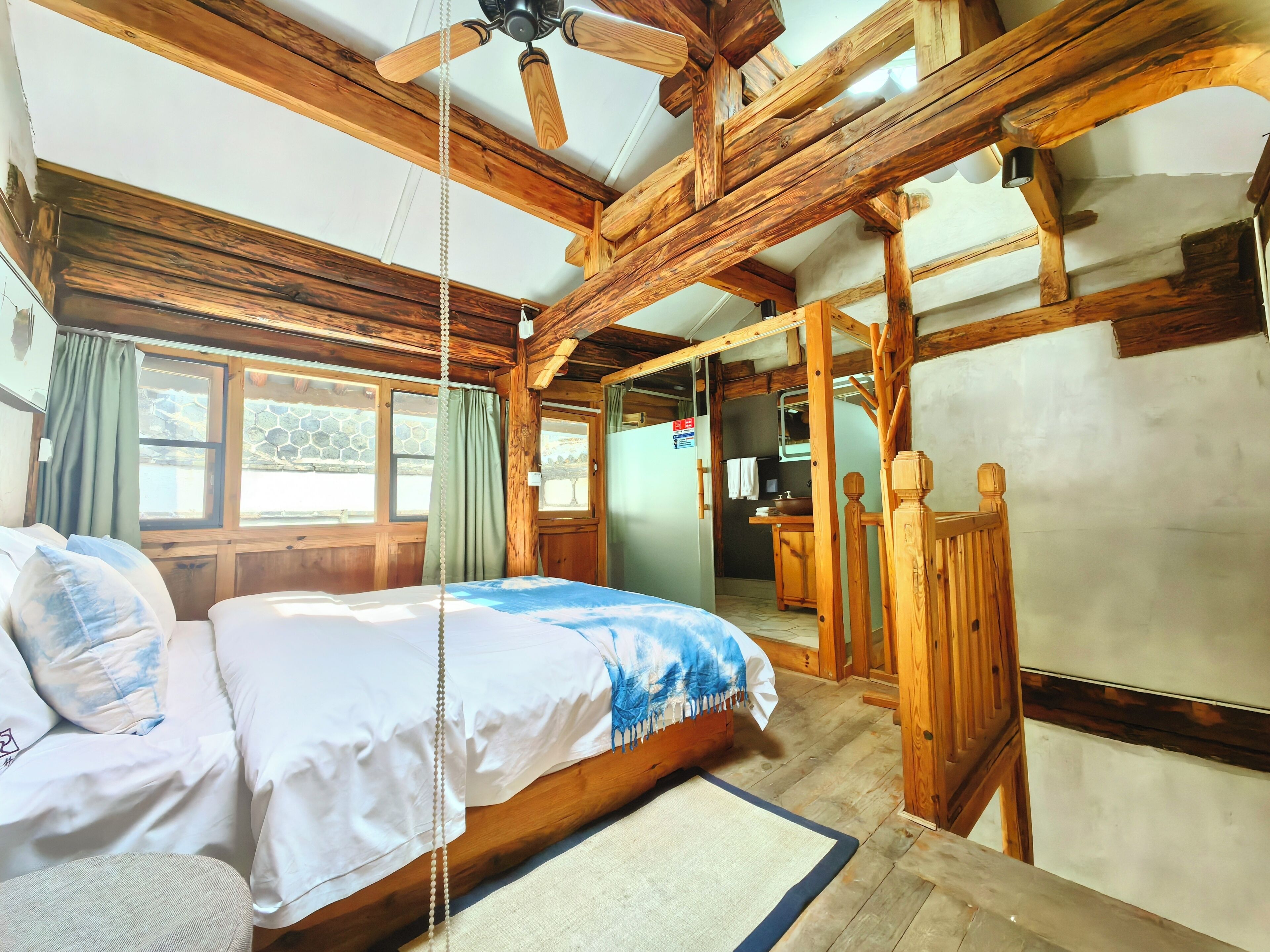 Una habitación tipo cabaña de madera con cama, ventilador de techo de madera, baño con espejo y vista al exterior a través de una ventana.