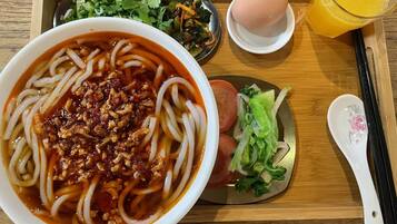 A meal tray with a bowl of noodles topped with a red sauce and minced meat, a small plate of sliced meat, a bowl of soup, a boiled egg, a glass of orange juice, and a spoon.
