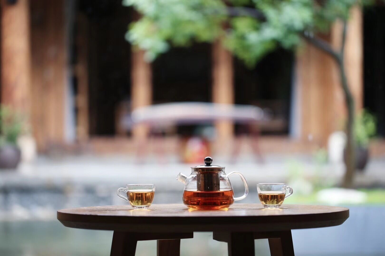 A table with a teapot and two cups of tea, set against a blurred background of a building and greenery.