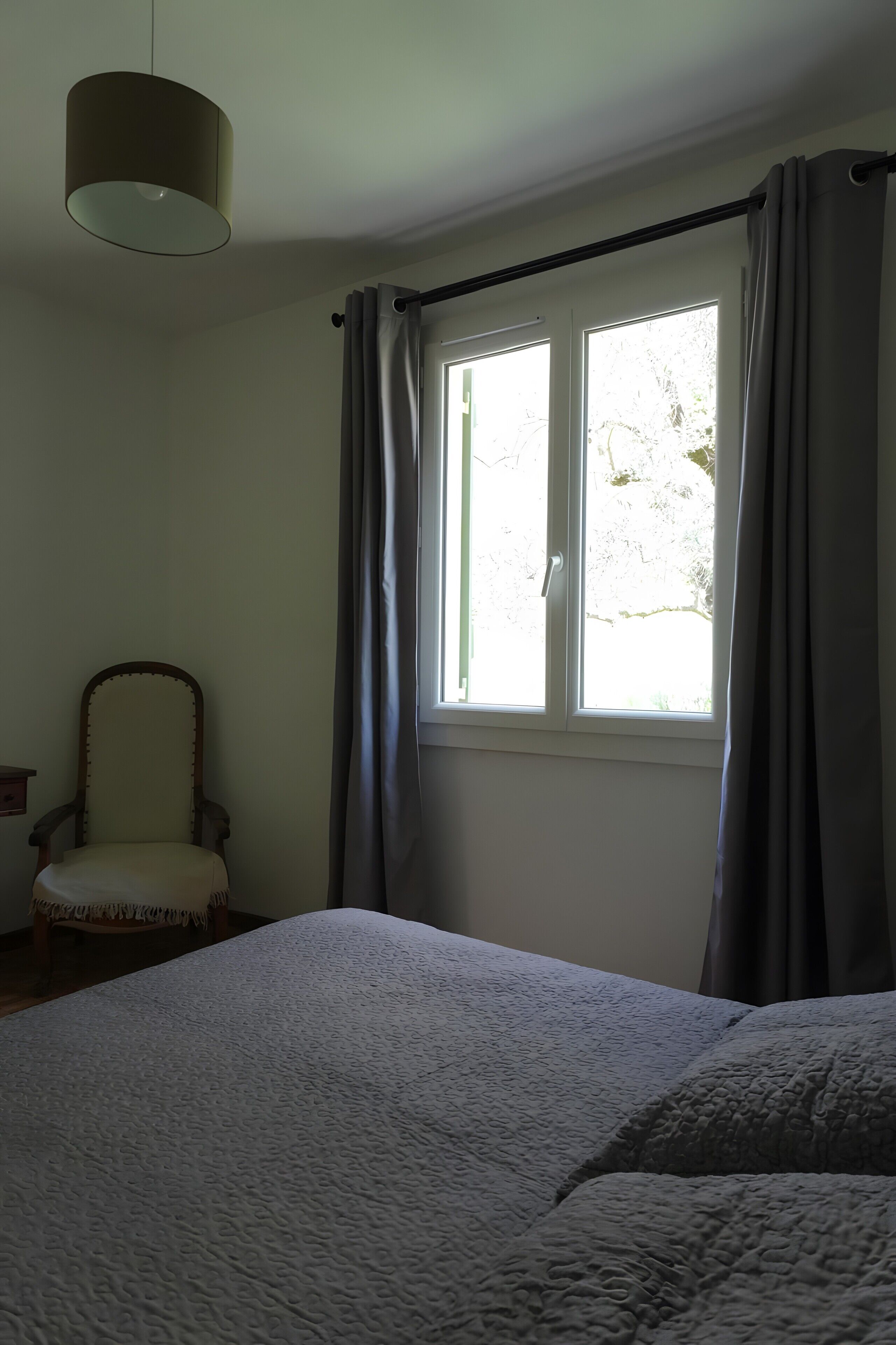 A bedroom with a bed, a chair, a window with curtains, and a hanging light fixture.