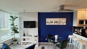 Ein modernes Wohnzimmer mit Flachbildfernseher, einem Essbereich mit Glastisch und einer Küche mit weißen Schränken.