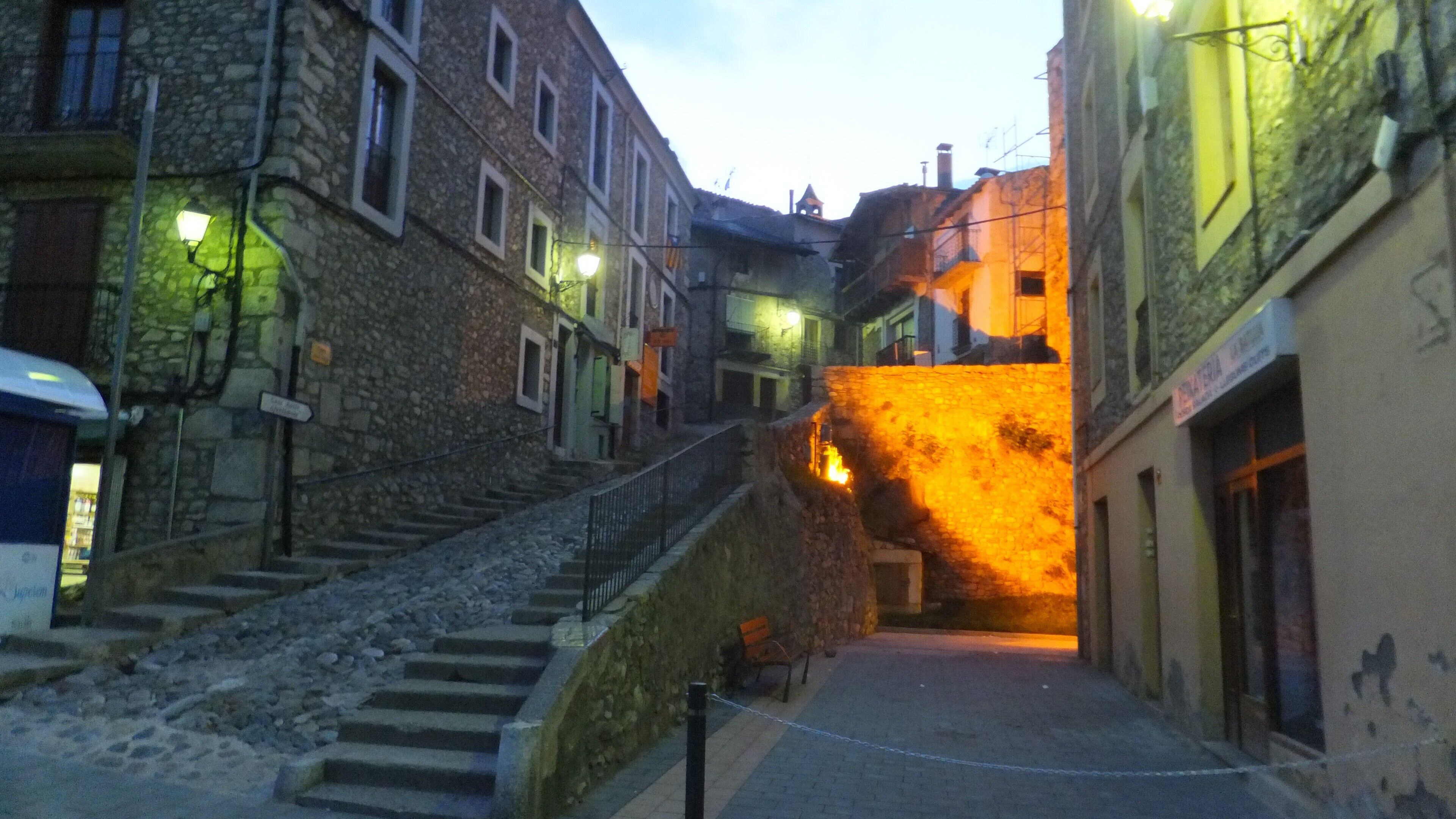 A narrow street with stone buildings, a staircase, and a brightly lit entrance.