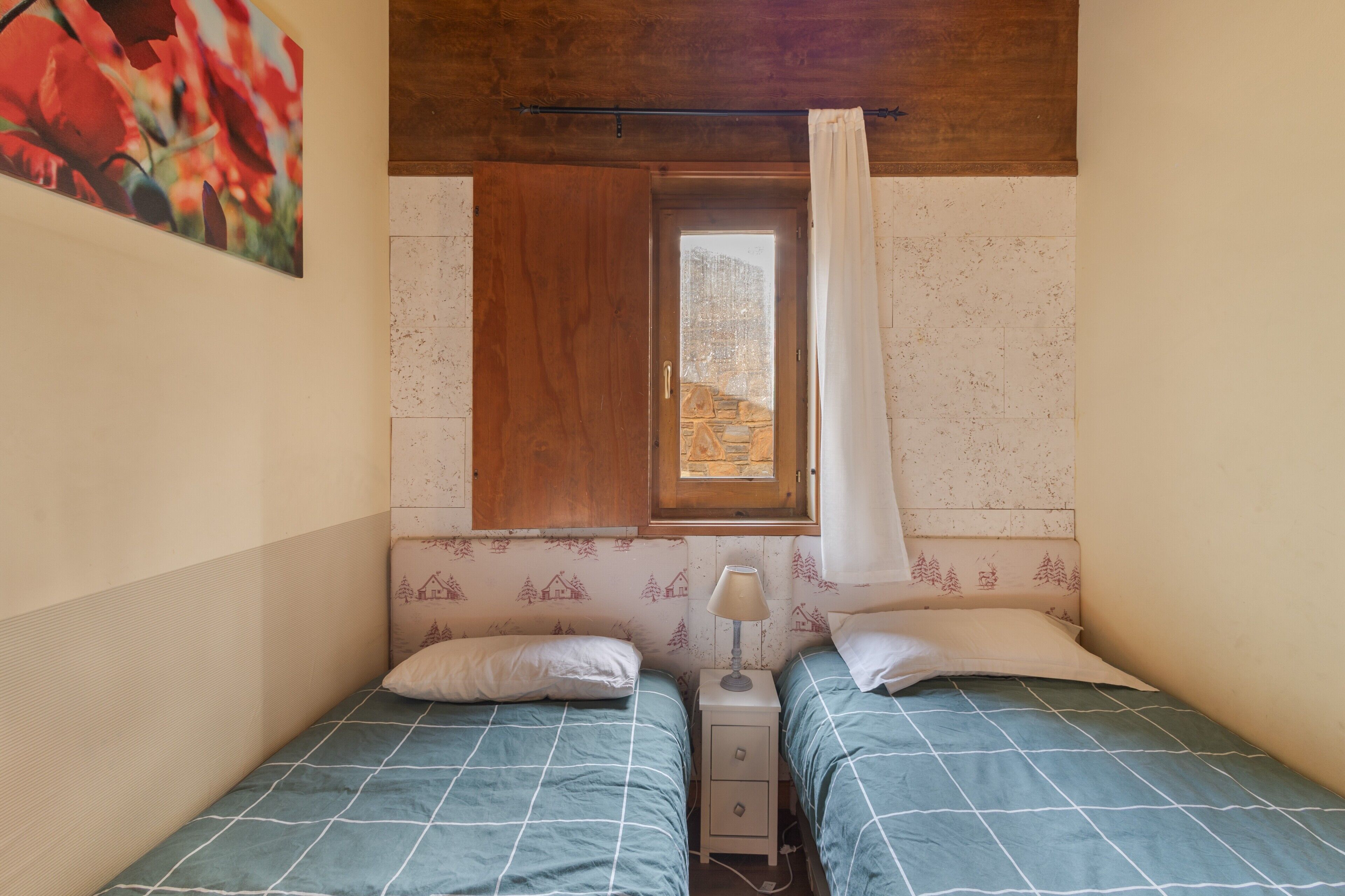 Un dormitorio con dos camas, una ventana con cortina, una mesita de noche con lámpara y una pared con estampado floral.