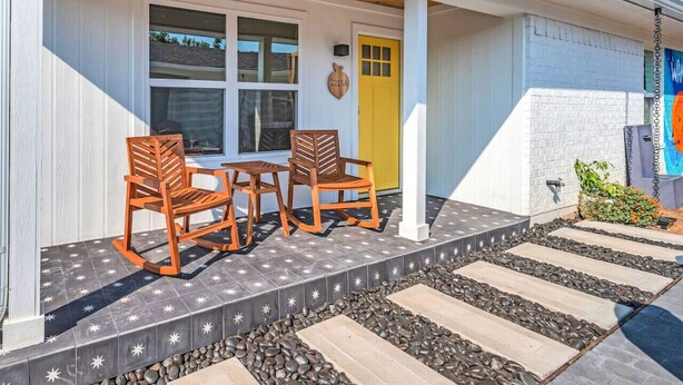A building with a porch featuring two rocking chairs and a table, a yellow door, and a small garden area with rocks and plants.