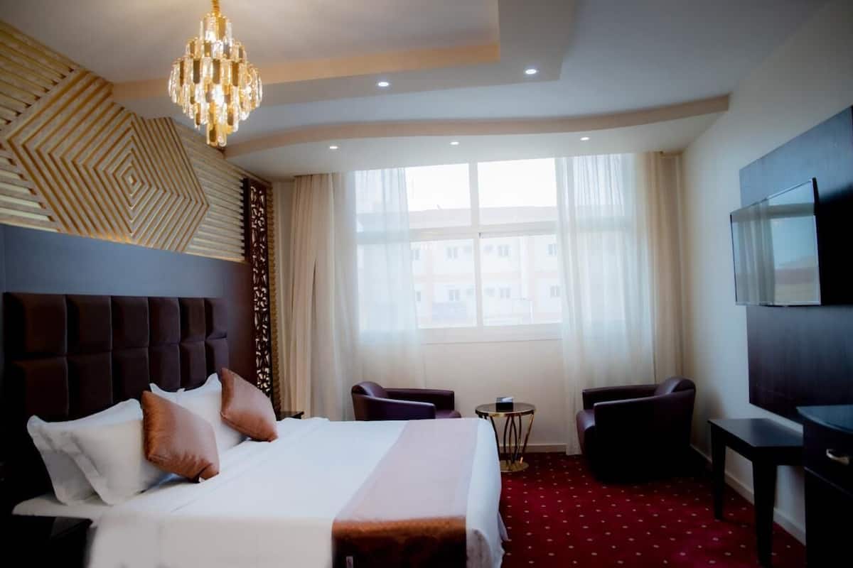 Habitación de hotel con una cama grande, una araña, dos sillones, una mesita y un televisor en la pared.