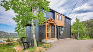 A modern two-story house with a wooden porch, a stone pathway leading to the entrance, and a view of a forested hillside.