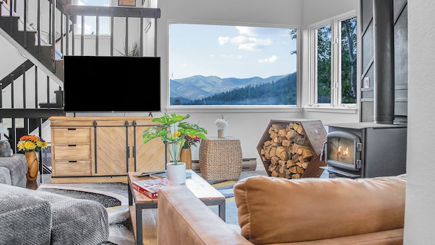 A modern living room with a large flat-screen TV, a fireplace, a sofa, and a view of mountains through large windows.