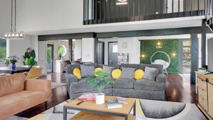 A spacious living area with a grey sofa, a coffee table, and a dining table with chairs. There is a staircase leading to an upper level.