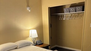 Un lit bien fait, avec une couette blanche et un plaid gris, une lampe de chevet, une table de chevet et un dressing où sont rangés des vêtements sur des cintres et des boîtes de rangement.