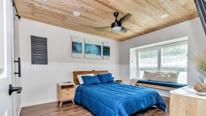 A bedroom with a wooden ceiling, a bed with blue bedding, a nightstand, a bedside table, and a window with blinds.