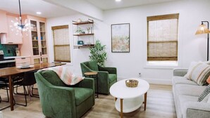 Een moderne woonkamer met groene fauteuils, een ronde salontafel en een grijze bank. Er is een eethoek met een houten tafel en stoelen, plus een keuken met een fornuis en kastjes.
