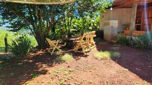 An outdoor seating area with wooden chairs and a table, surrounded by greenery and a building with a wooden exterior. - Fully equipped cabins, on the jungle route on the way to the Moconá Falls. (El Soberbio)