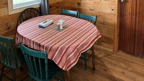 A wooden cabin with a dining table covered by a striped tablecloth, chairs, a hanging light fixture, a framed picture on the wall, and a window with curtains. - Seabreeze Retreat-Cozy waterfront  3-bedroom, one minute from PEI National Park (York)
