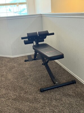 A corner of a room with a black exercise bench against a wall, carpeted floor, and a window with blinds. - Contemporary family-friendly 4-bedroom home in Killeen, located near Fort Hood! (Killeen)