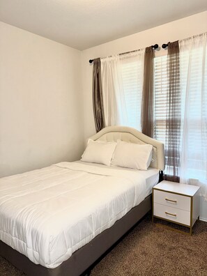 A neatly made bed with white bedding, a bedside table with a drawer, and a window with sheer curtains. - Contemporary family-friendly 4-bedroom home in Killeen, located near Fort Hood! (Killeen)