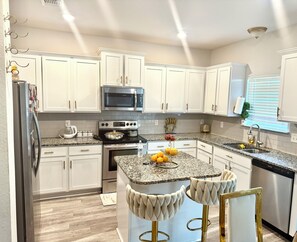 Una cocina moderna con armarios blancos, electrodomésticos de acero inoxidable, encimeras de granito y una isla central con taburetes de bar.