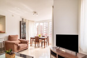 A modern living room with a sofa, a dining table with chairs, a flat-screen TV, and a large window with a city view.