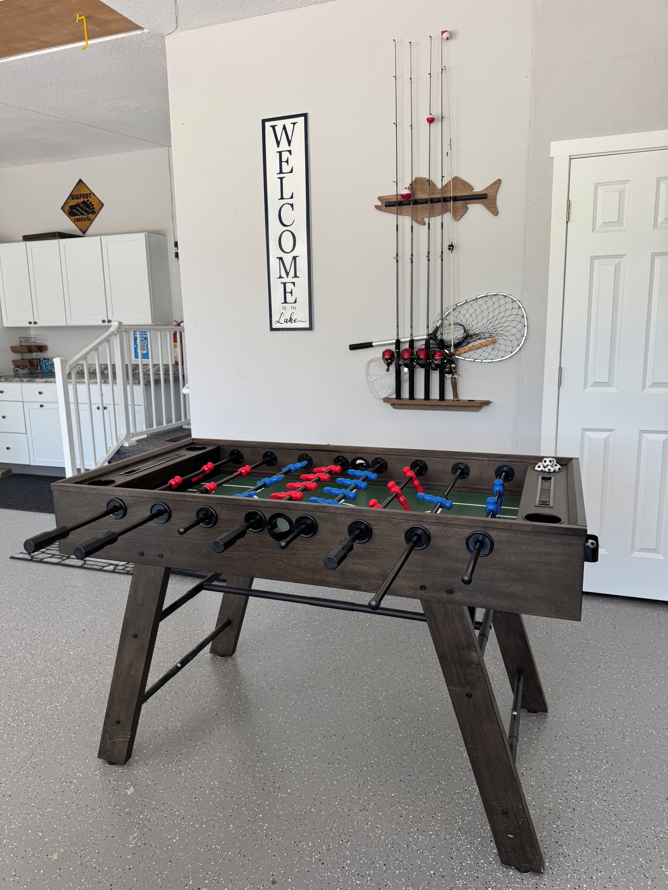 A foosball table in a room with a 'WELCOME' sign, fishing rods, and a door.