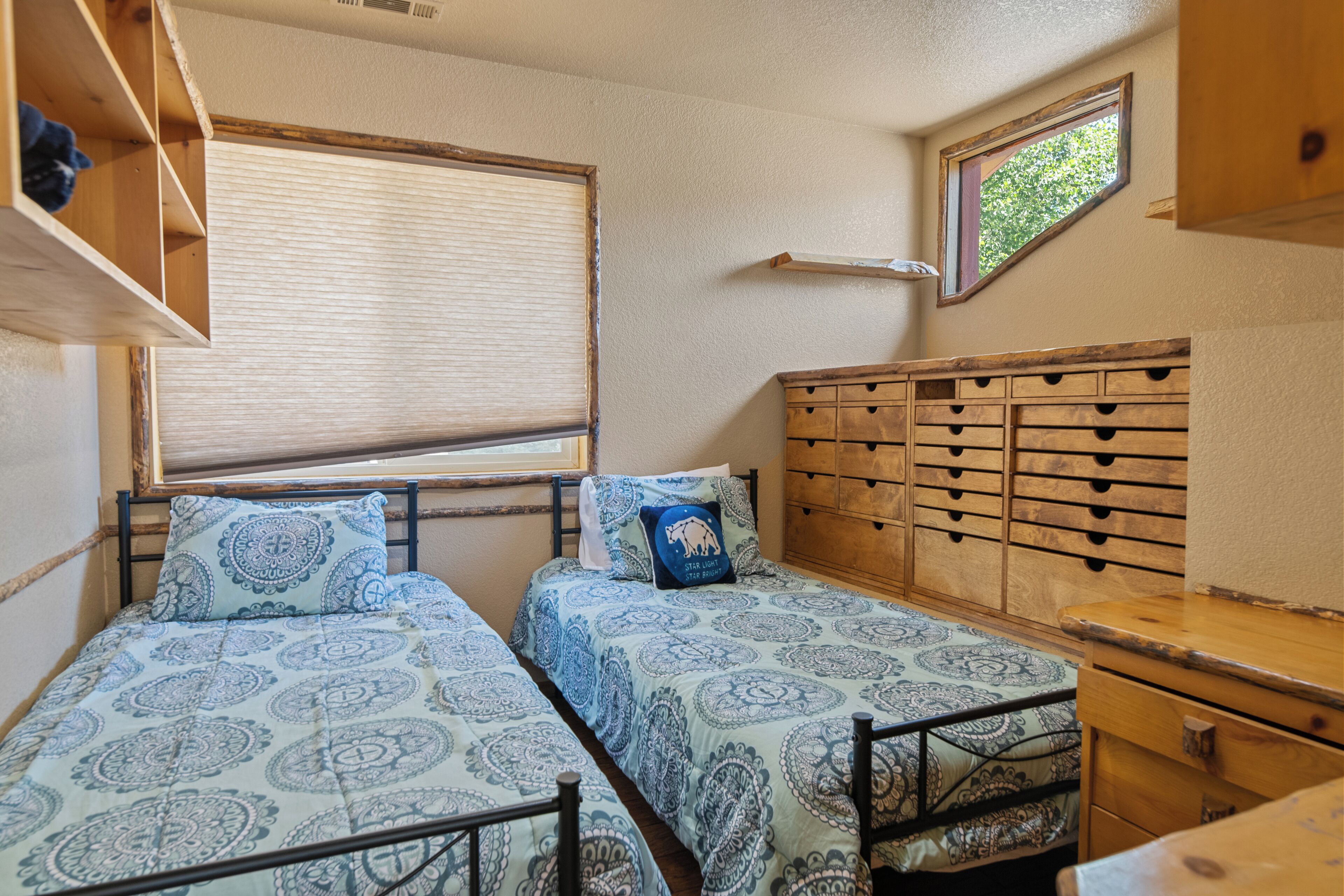 Una habitación compacta con dos camas individuales, un aparador de madera, una ventana con persianas y un tragaluz pequeño.