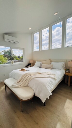Un dormitorio amplio con una cama grande, un banco y una ventana que ofrece vistas a zonas verdes y un edificio.