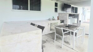 Un espace cuisine extérieur abrité, doté d’un plan de travail en marbre, d’une table à manger avec des chaises, d’un évier encastré et d’un téléviseur fixé au mur.
