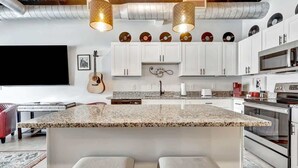 Una cocina moderna con armarios blancos, encimera de granito y una isla central con asientos. Hay luces colgantes, una guitarra y un reloj en la pared.