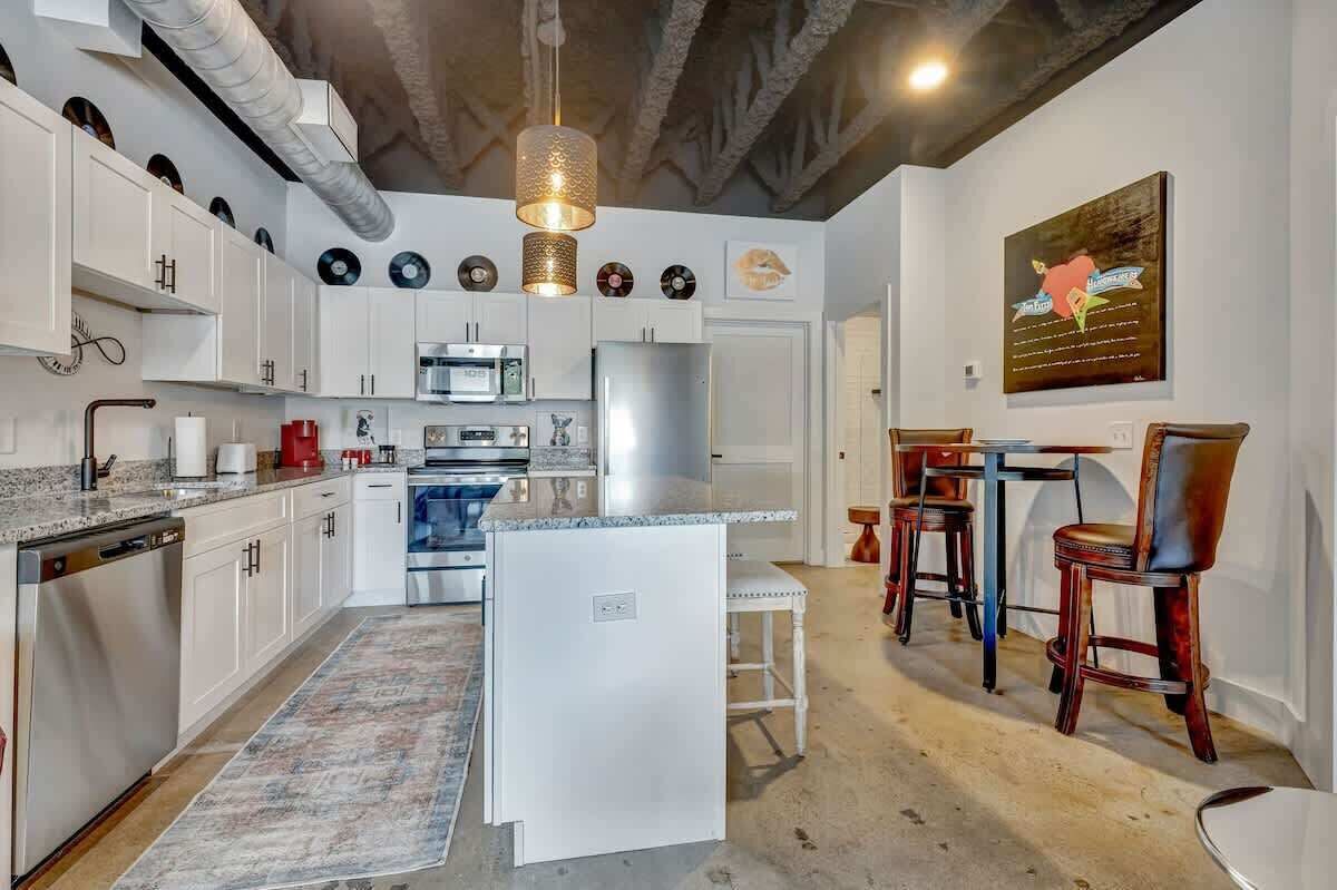 Una cocina moderna con armarios blancos, microondas integrado y refrigerador. Hay un comedor con sillas de madera y una mesa.