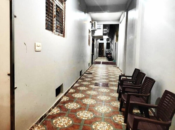 Un couloir étroit, dont le sol est recouvert de carreaux décoratifs, une rangée de chaises en plastique d’un côté, et une moto garée au bout.