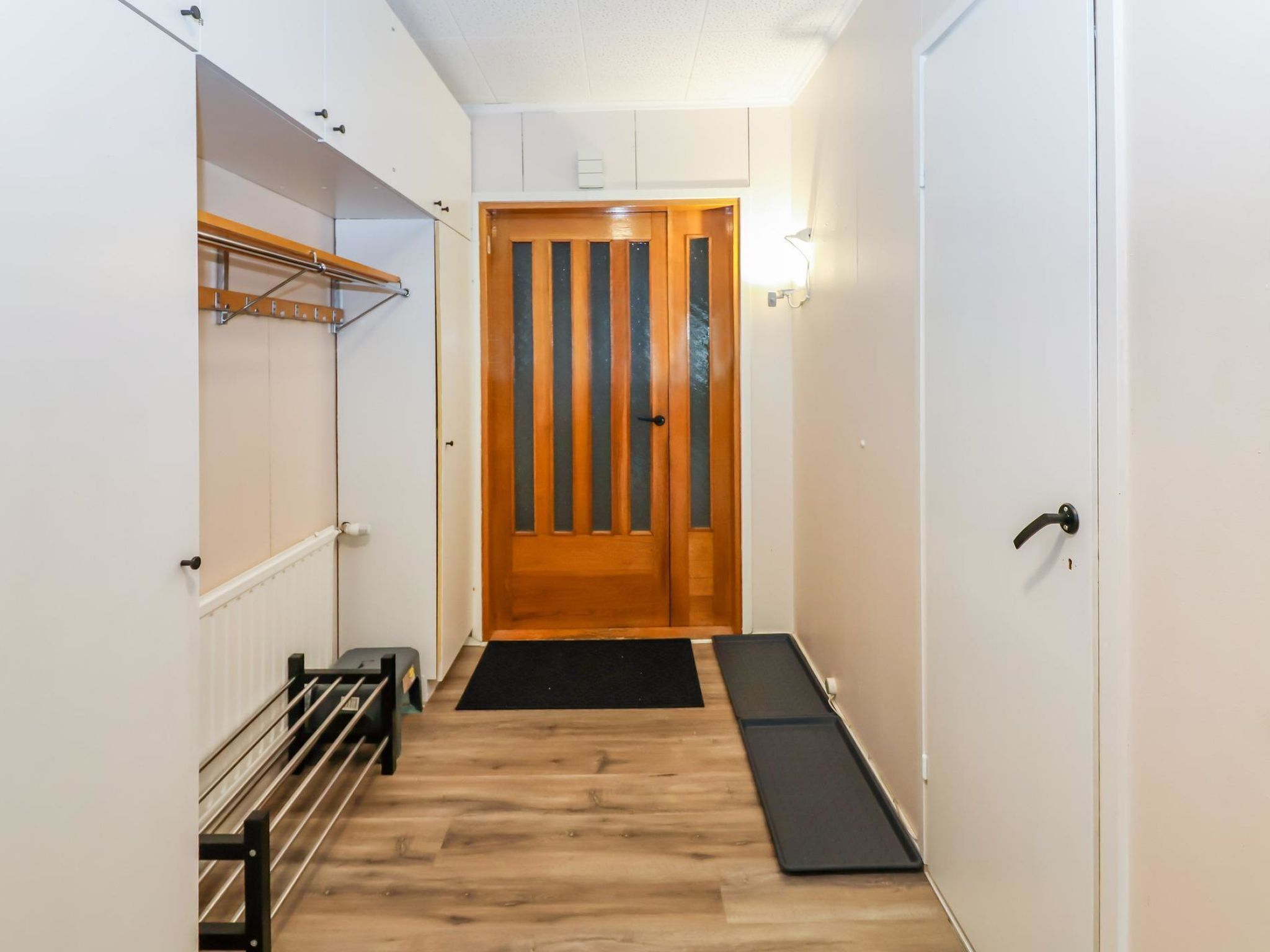 A narrow hallway with a wooden door at the end, a black mat at the entrance, and a metal rack on the left wall.