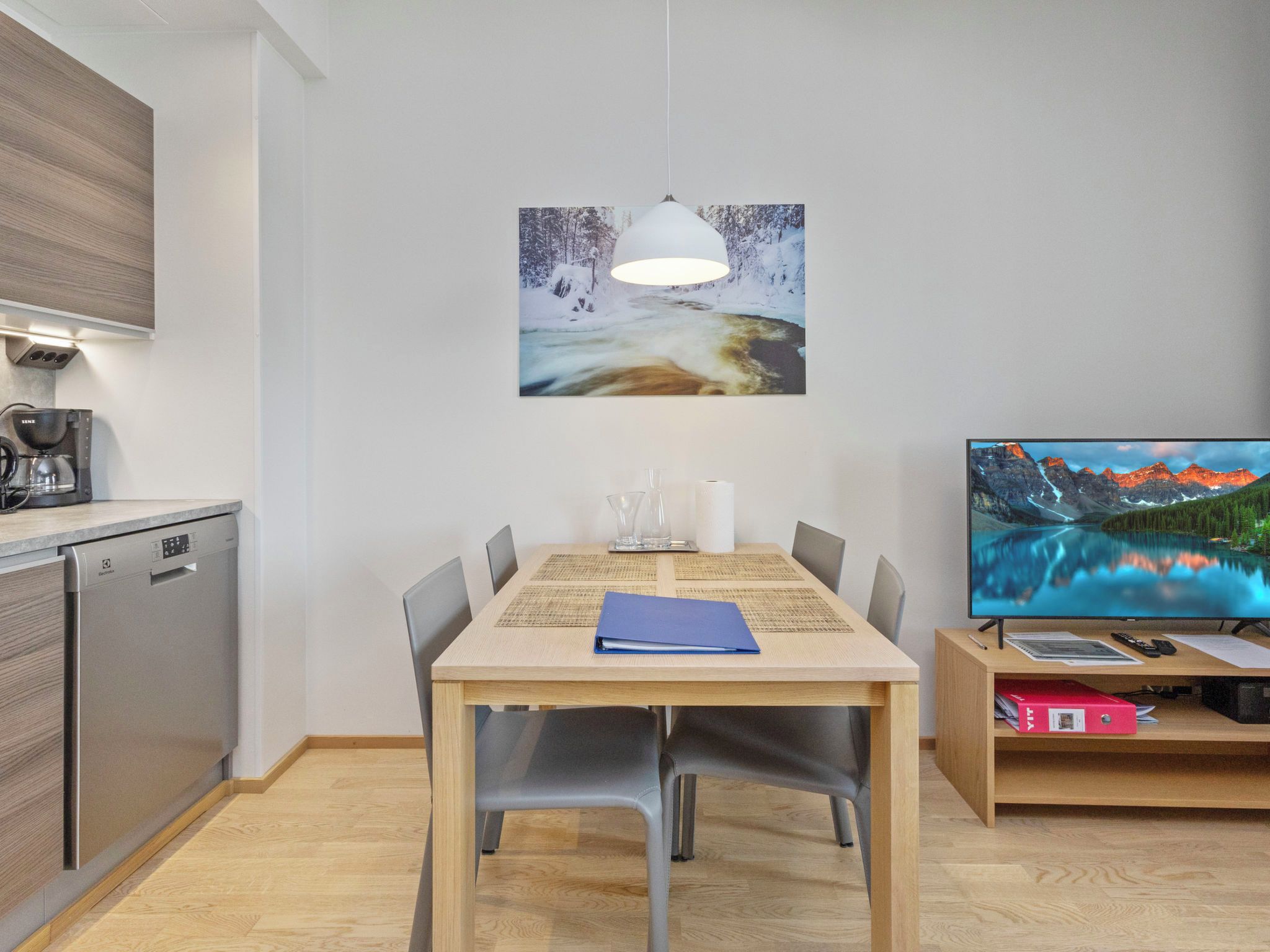 A modern kitchen with a coffee machine, a dining table with chairs, a painting, and a television.