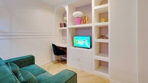 A living room with a green sofa, a black chair, a TV on a white cabinet, and shelves with decorative items. - Cosy Appt - 1bdr/2p - Moulin Rouge / Montmartre (Paris)