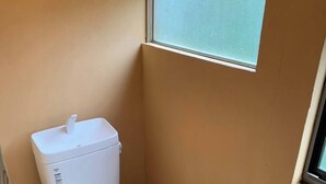 Een kleine badkamer met een wit toilet, een klein raam met een hor en een toiletrolhouder aan de muur.