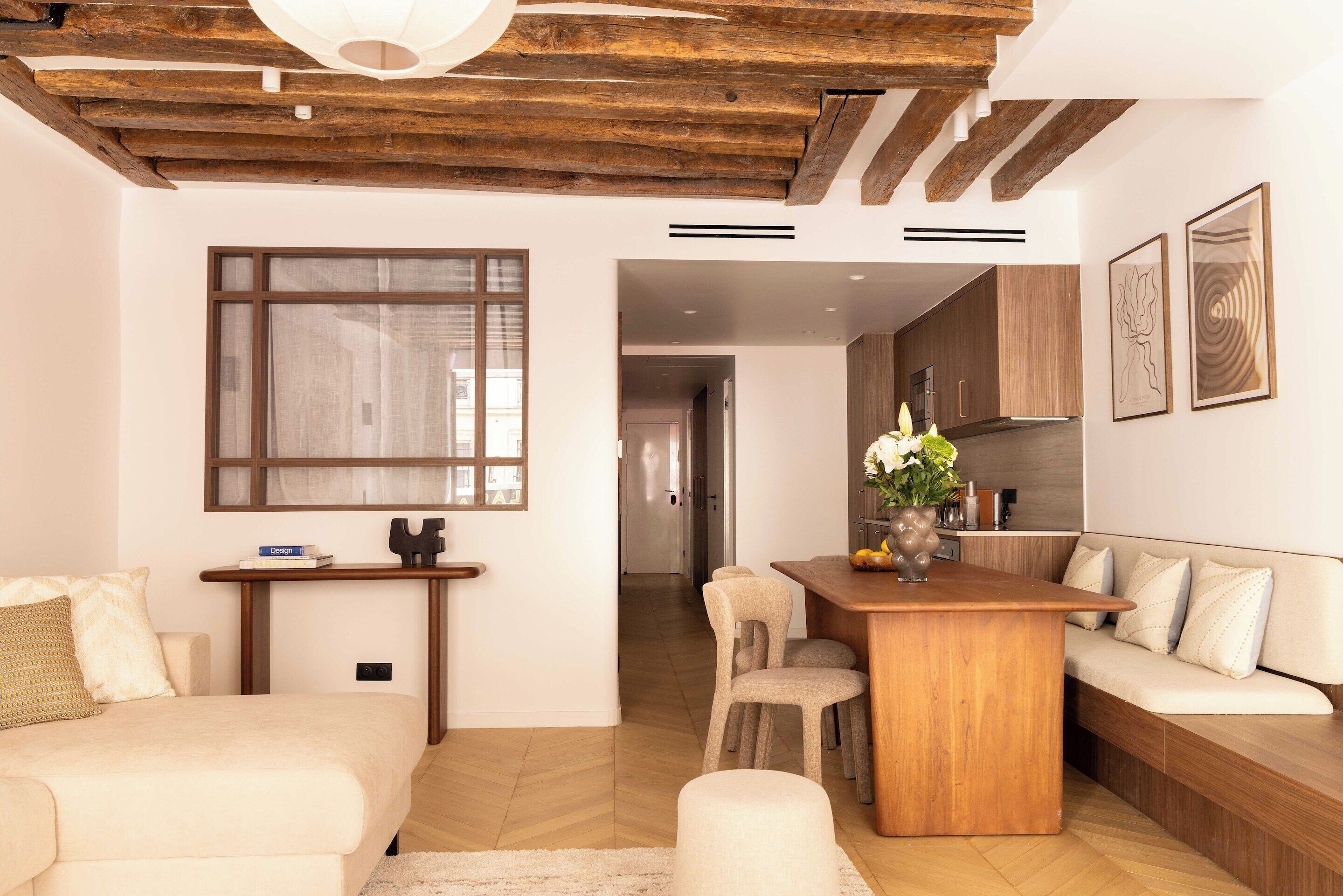 A modern living room with a wooden ceiling, a large window, a dining area with a wooden table and chairs, and a sofa with cushions.
