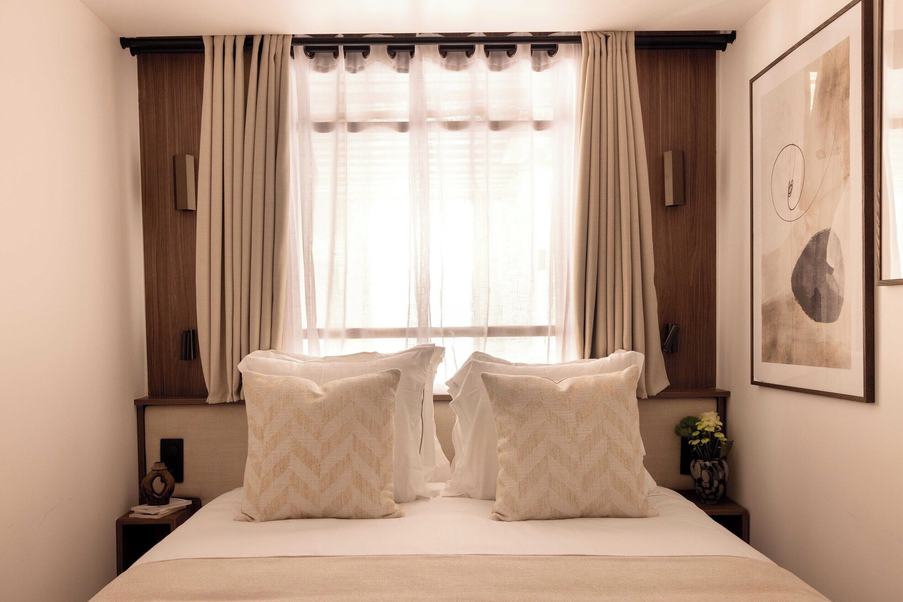 A bedroom with a large bed, two bedside tables, a window with curtains, and a wall-mounted artwork.