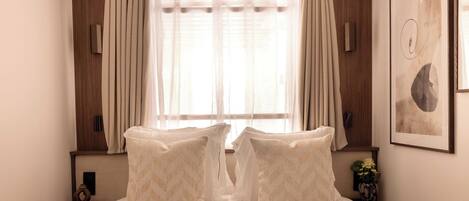 A bedroom with a large bed, two bedside tables, a window with curtains, and a wall-mounted artwork.