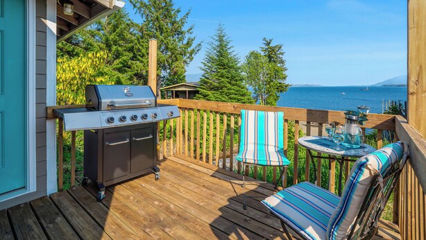 Une terrasse en bois avec un barbecue, une chaise à rayures et une table avec une carafe.