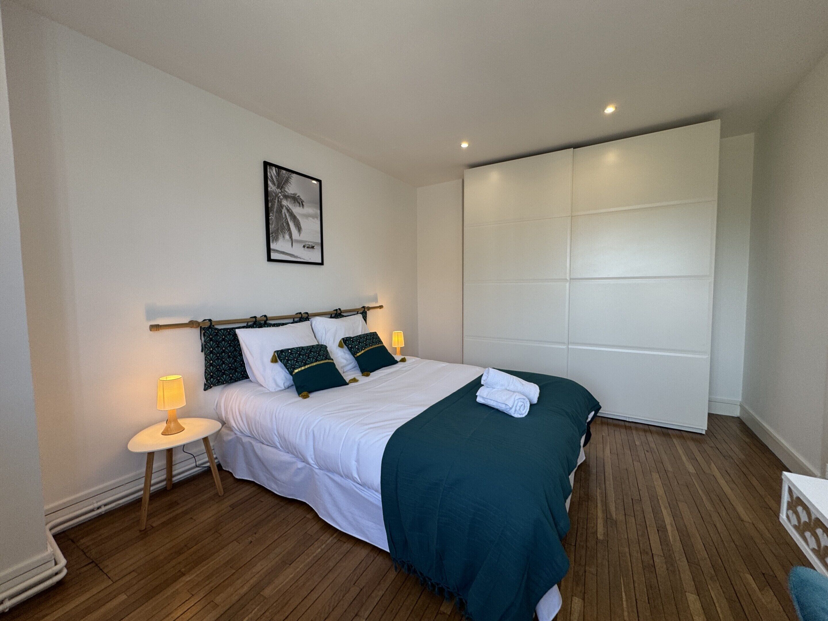 A neatly arranged bedroom with a bed, bedside tables, a wall-mounted artwork, and a large wardrobe.