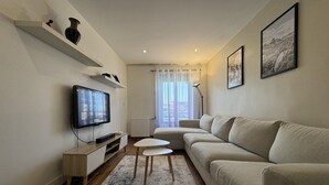 Een moderne woonkamer met een flatscreen televisie, een beige bank, een kleine witte tafel en een vloerkleed met een patroon.