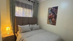 A bedroom with a bed, a nightstand, a lamp, a window with curtains, and a wall-mounted painting. - Kisasi Delight (Kampala)
