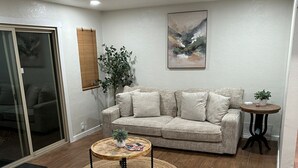 Sala de estar con sofá, mesa de centro y una silla. Hay una puerta que da a un balcón, una planta y un cuadro en la pared.